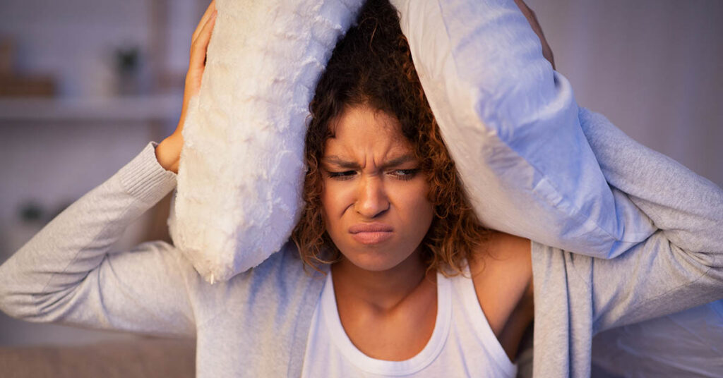 Woman covering ears with pillows due to loud noise from neighbors, illustrating HOA noise complaints